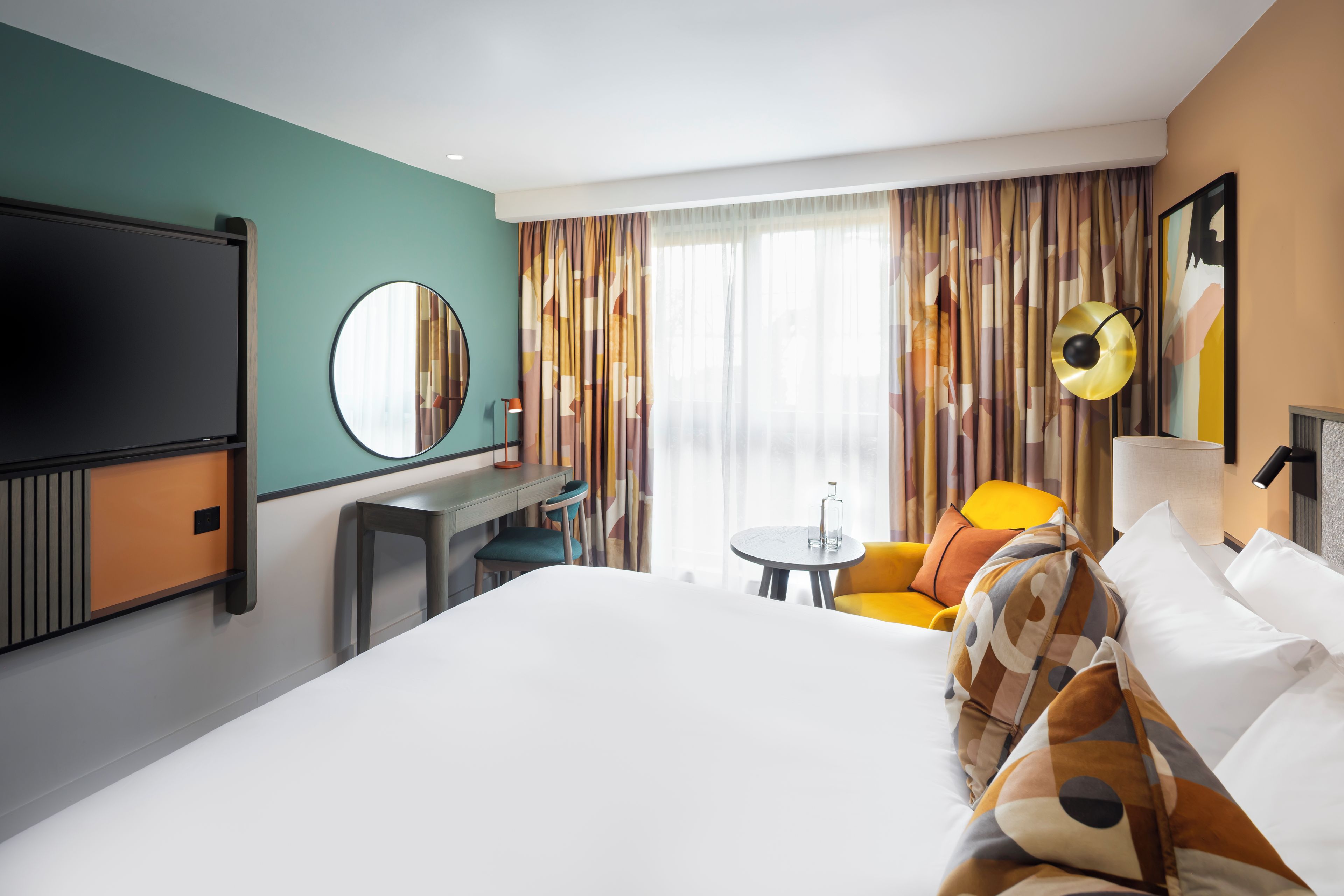 A hotel room with a bed, a desk, a chair, a round mirror, a lamp, and a window with curtains.