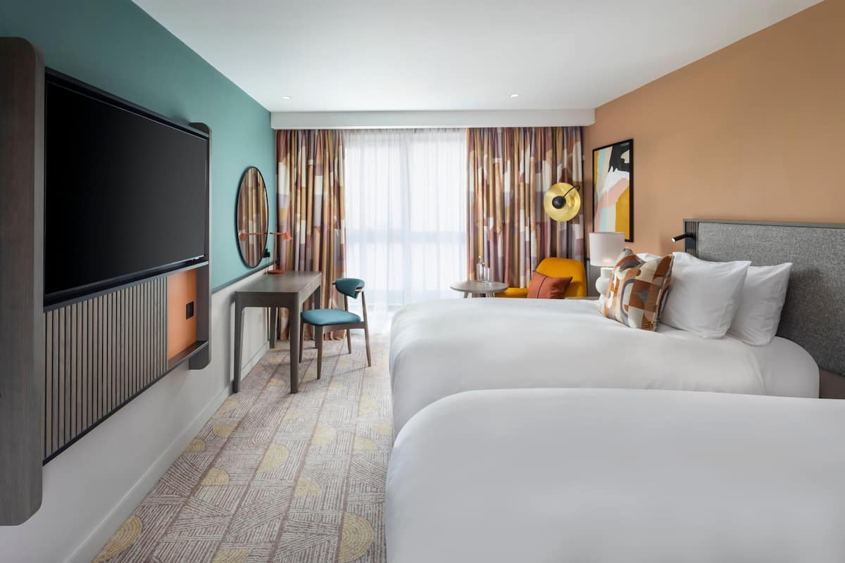 A hotel room with two beds, a TV, a desk, and a large window with curtains.