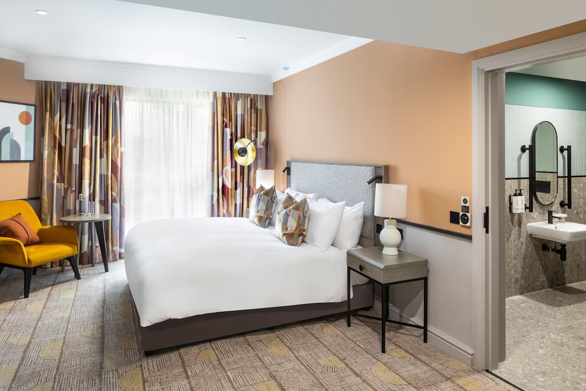 A hotel room with a large bed, a bedside table with a lamp, a chair, a small table, and a bathroom with a sink and mirror.