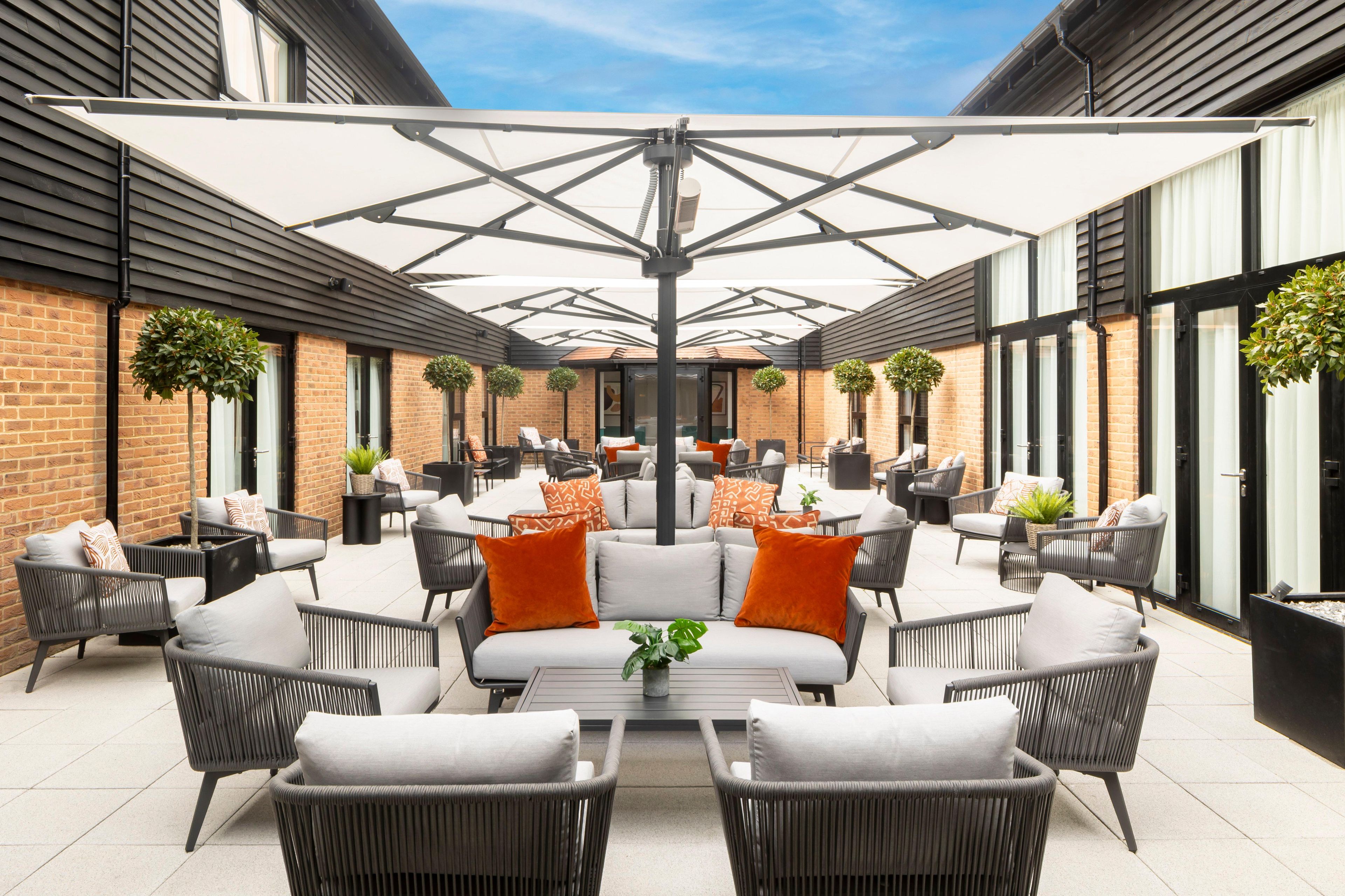 A covered outdoor seating area with wicker furniture and a central table, surrounded by brick walls and large windows.