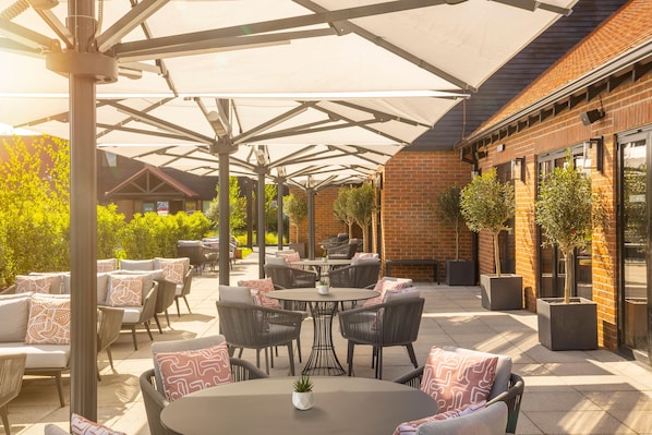 An outdoor patio area with round tables and chairs, a brick building, and potted plants. - Voco Fareham-solent (Fareham)
