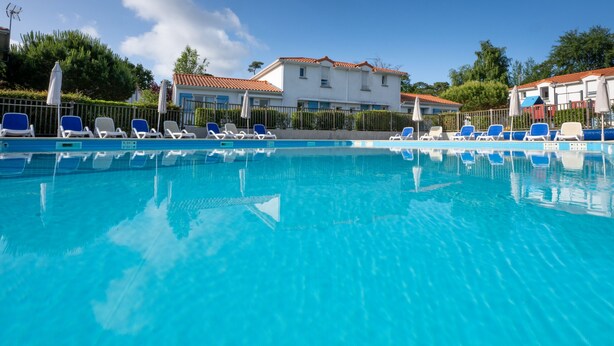 Una piscina con sillones de descanso y sombrillas, rodeada de edificios con tejados de tejas rojas.