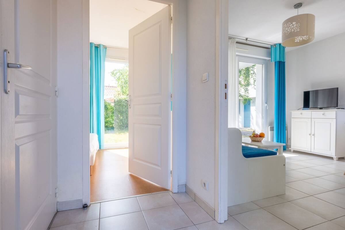 Une pièce lumineuse avec une porte blanche, un petit coin salon avec un coussin bleu, un téléviseur et une fenêtre ornée de rideaux bleus.