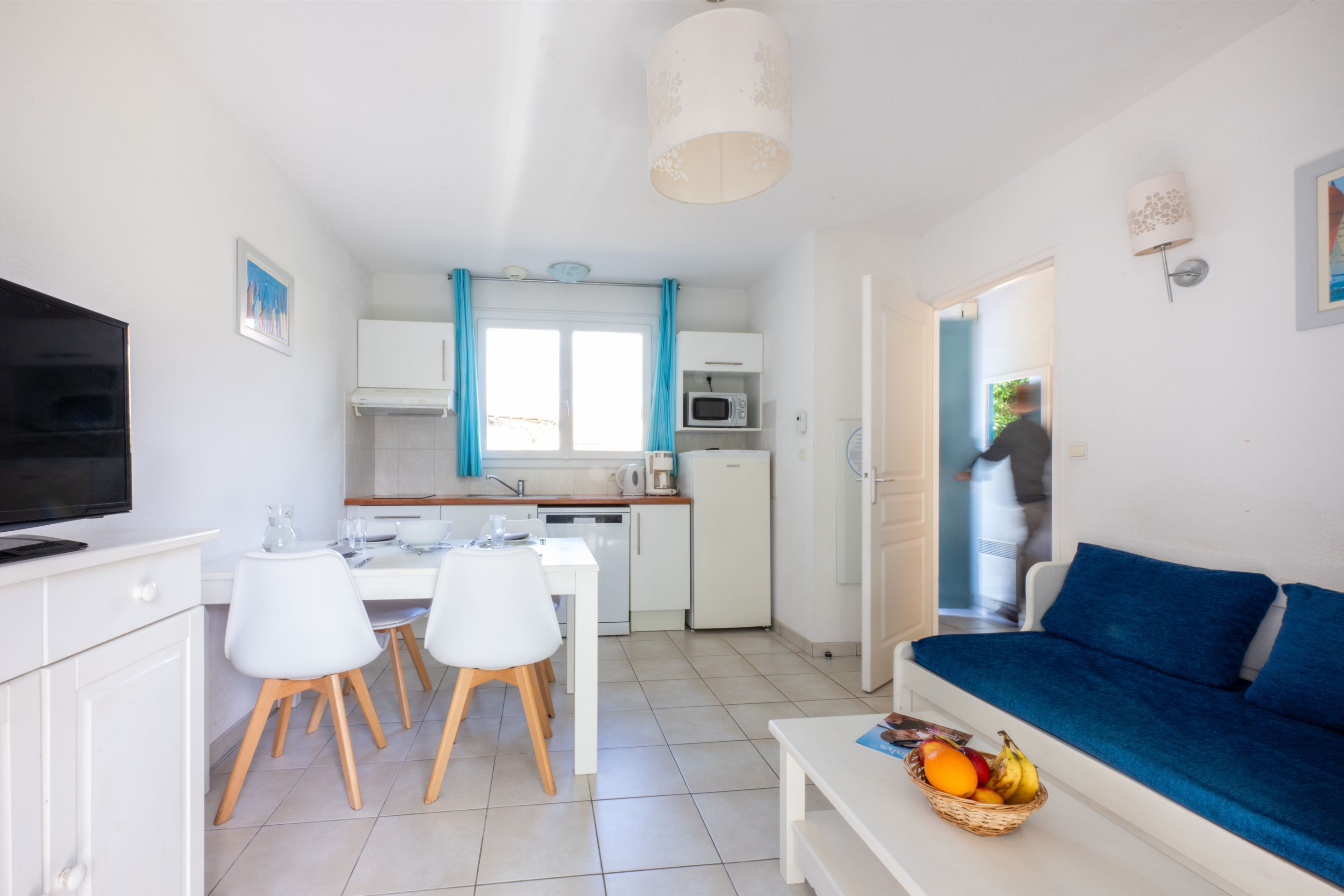 A modern living room with a blue sofa, a dining area with white chairs, and a kitchen with white cabinets and a microwave.