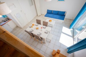 A modern, well-lit room with a dining table set for four, a blue sofa, and a kitchen area with a sink and cabinets. - Le Domaine de L'océan (Saint-Brevin-les-Pins)