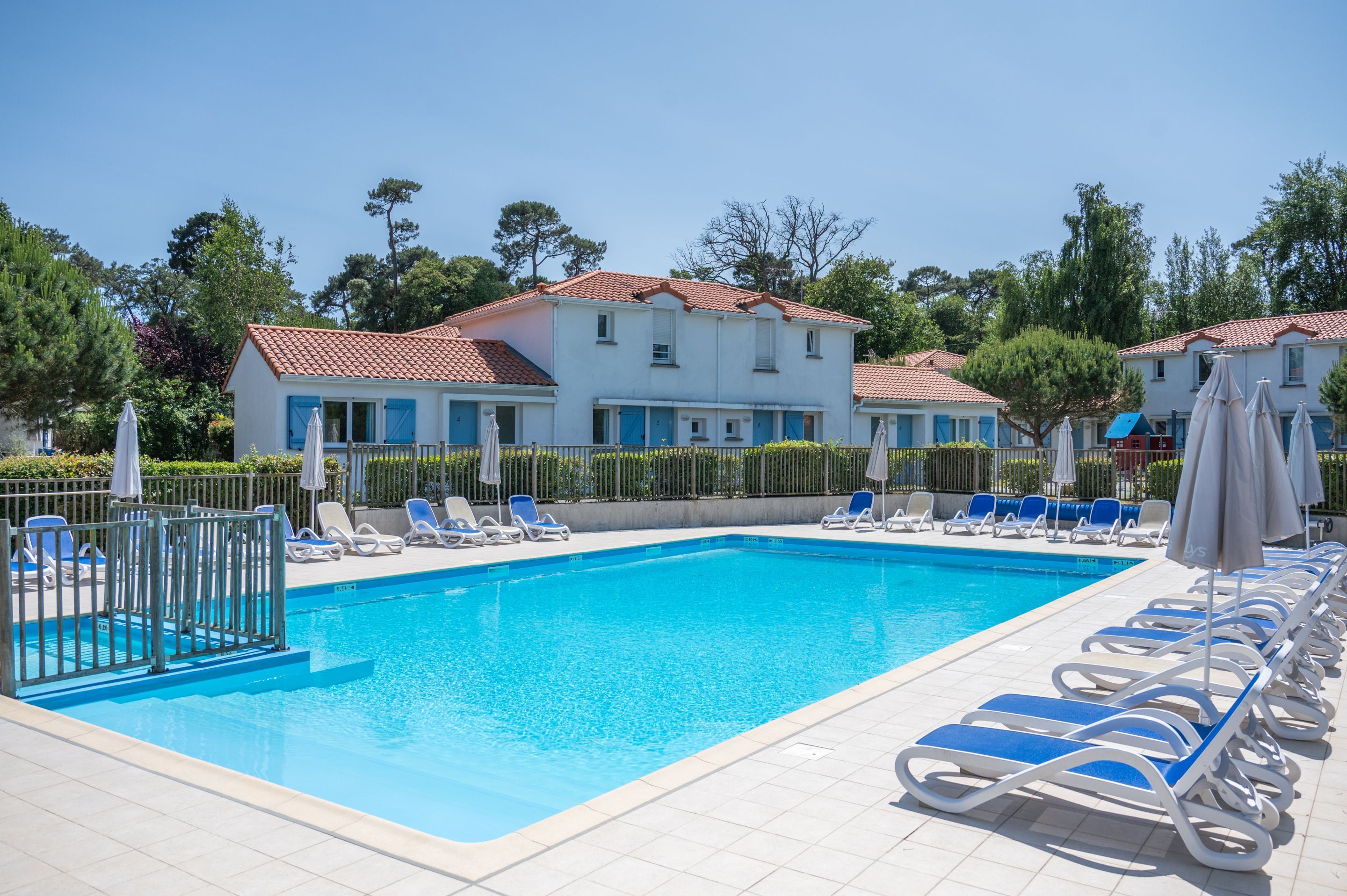 A swimming pool with blue lounge chairs and umbrellas, surrounded by a white building with a red roof.