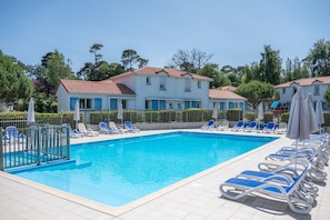 A swimming pool with blue lounge chairs and umbrellas, surrounded by a white building with a red roof. - Le Domaine de L'océan (Saint-Brevin-les-Pins)