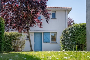 Un bâtiment à deux étages avec une porte et des fenêtres bleues, entouré de verdure et sous un ciel dégagé.
