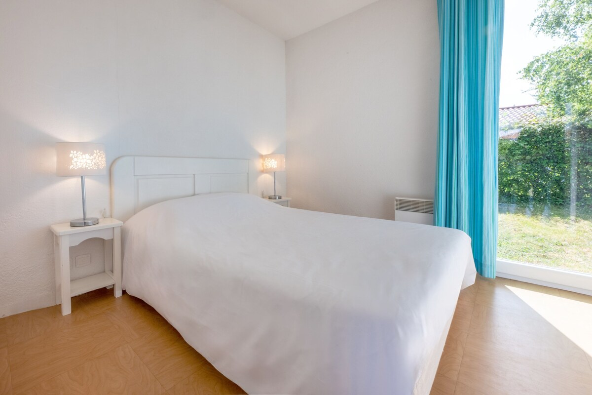 A bedroom with a large bed, two bedside tables with lamps, a window with curtains, and a view of greenery outside.