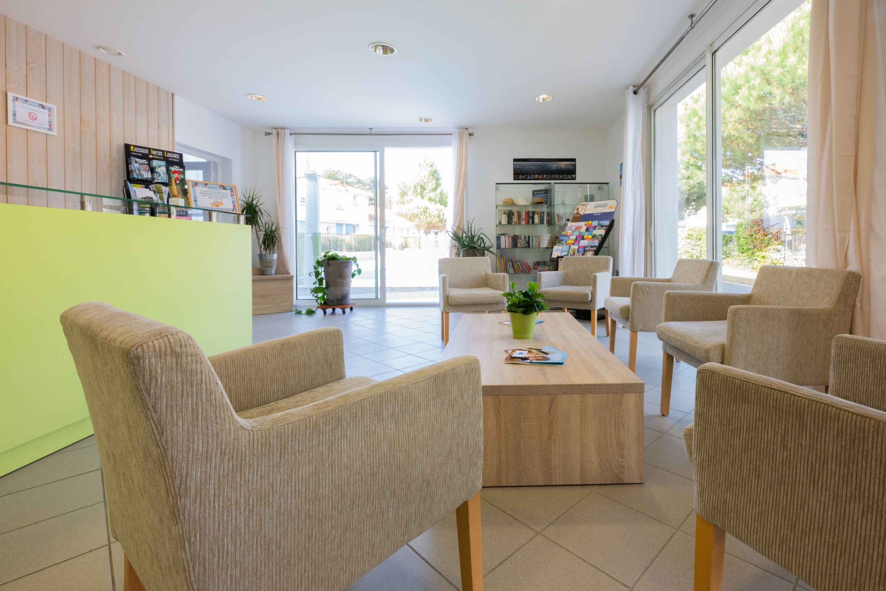 A reception area with a green counter, beige chairs, a wooden coffee table, and a glass door leading outside.