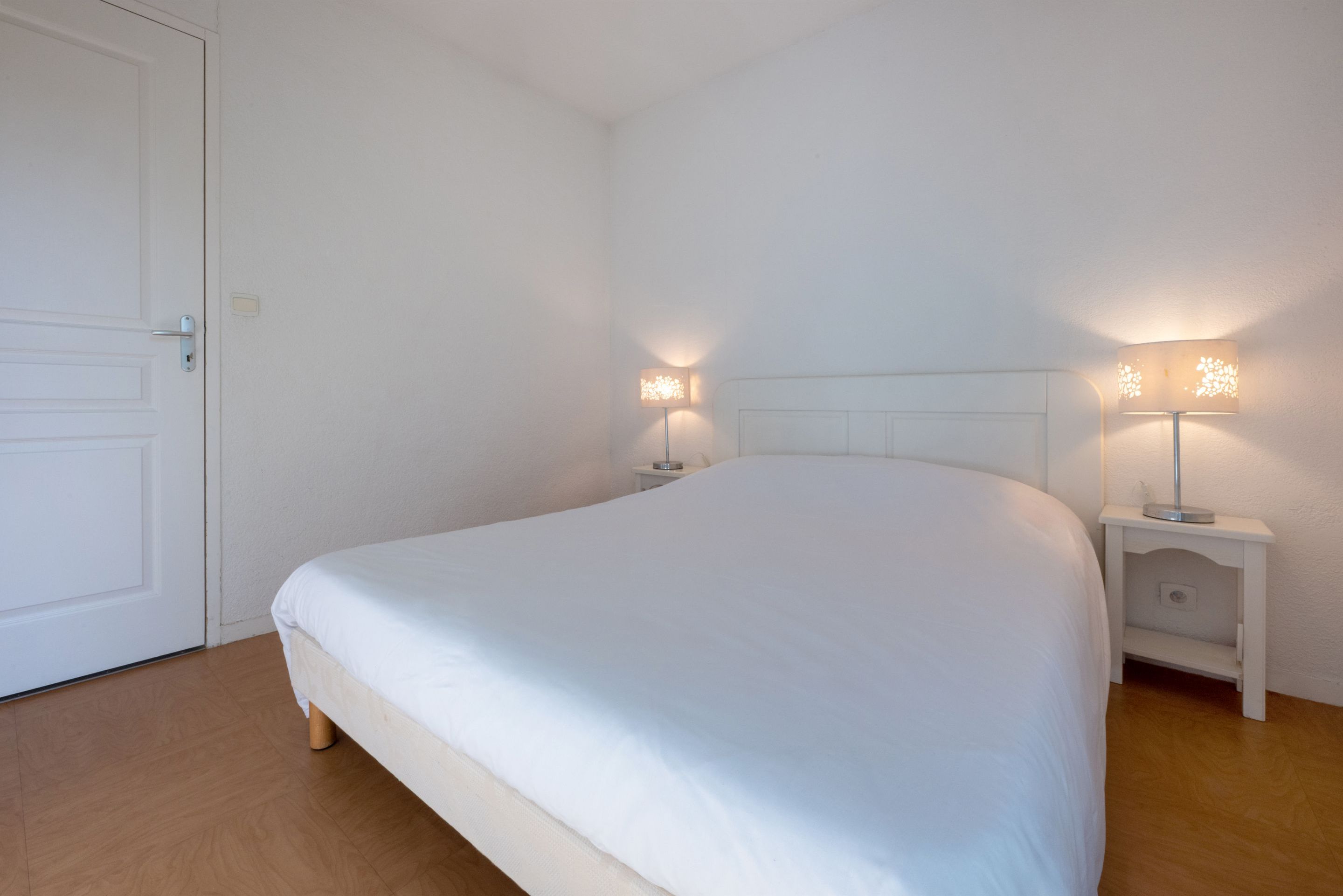 A neatly made bed in a room with white walls and a wooden floor. There are two bedside tables with lamps.