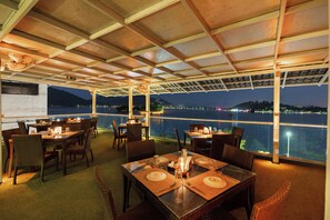 Een restaurant met gedekte tafels, met uitzicht op een waterpartij 's nachts.
