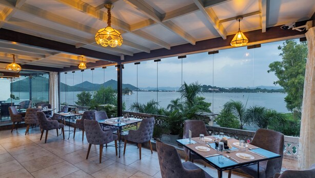 A restaurant with tables and chairs, large windows, and a view of a body of water. - Panna Villas Udaipur (Udaipur)