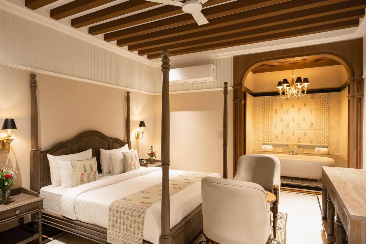 A four-poster bed with a nightstand, a chair, a desk, and a bathroom with a bathtub and a chandelier.