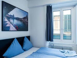 Una habitación de hotel con cama, ventana con cortinas, radiador y un cuadro en la pared.