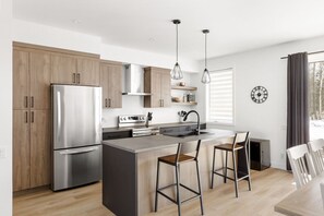 A modern kitchen with wooden cabinets, stainless steel appliances, a central island with bar stools, pendant lighting, and a window with blinds. - Le Memph Orignal: 3 bedrooms pool hot tub lac Memphrémagog (Magog)