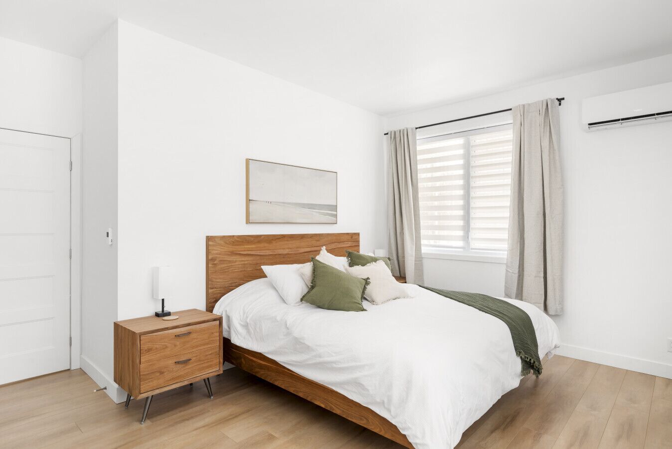 A bedroom with a wooden bed, white bedding, a bedside table with a lamp, and a window with curtains.