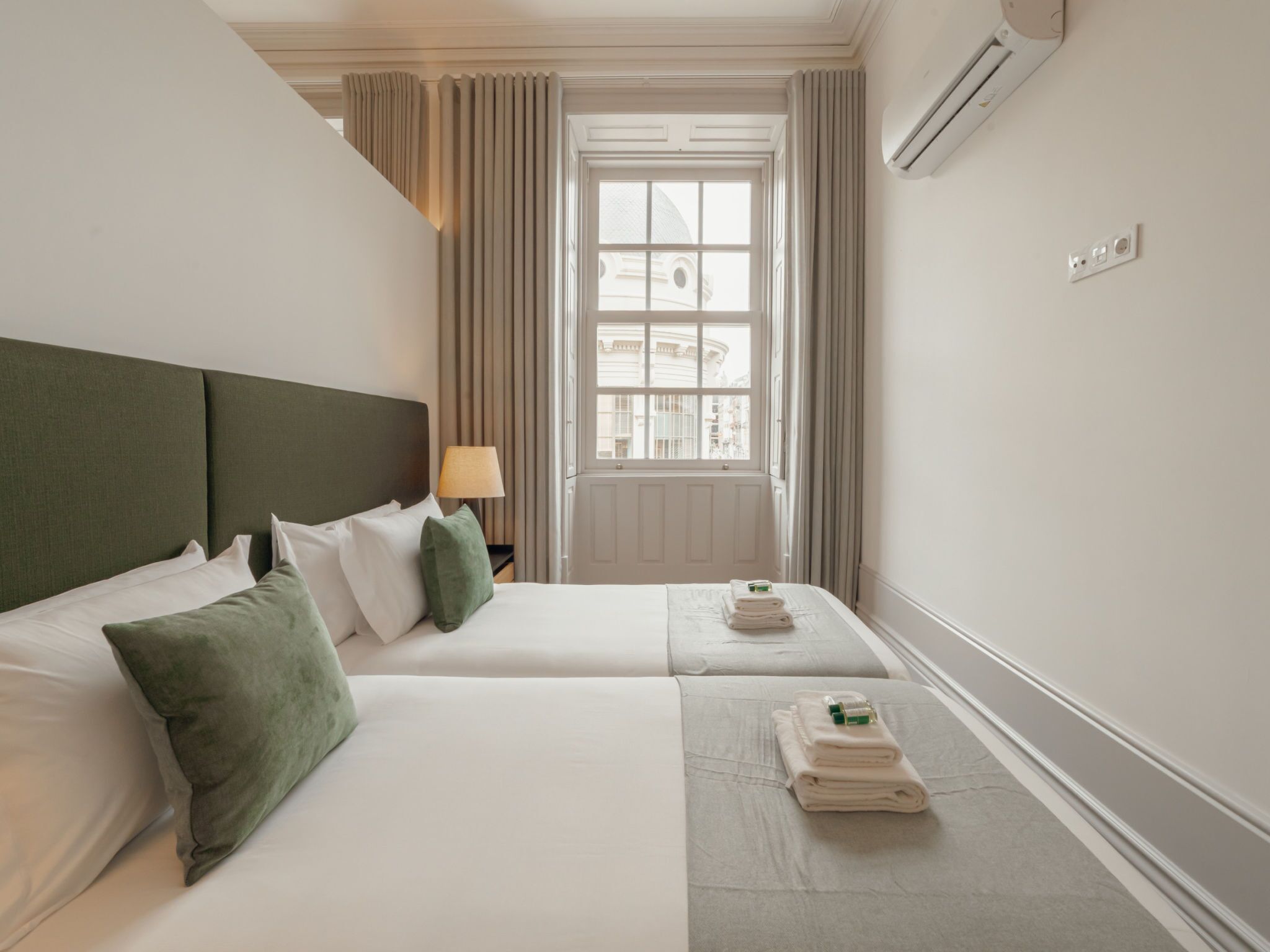 A hotel room with two beds, a window with curtains, a lamp, and an air conditioning unit.