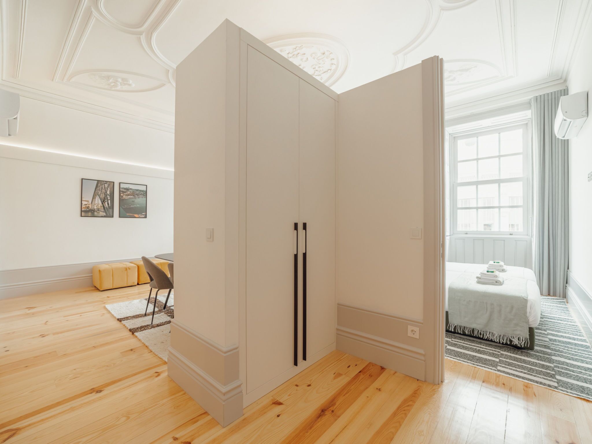 A room with a large wardrobe, a bed with a white bedspread, a window with curtains, and a view of a building.