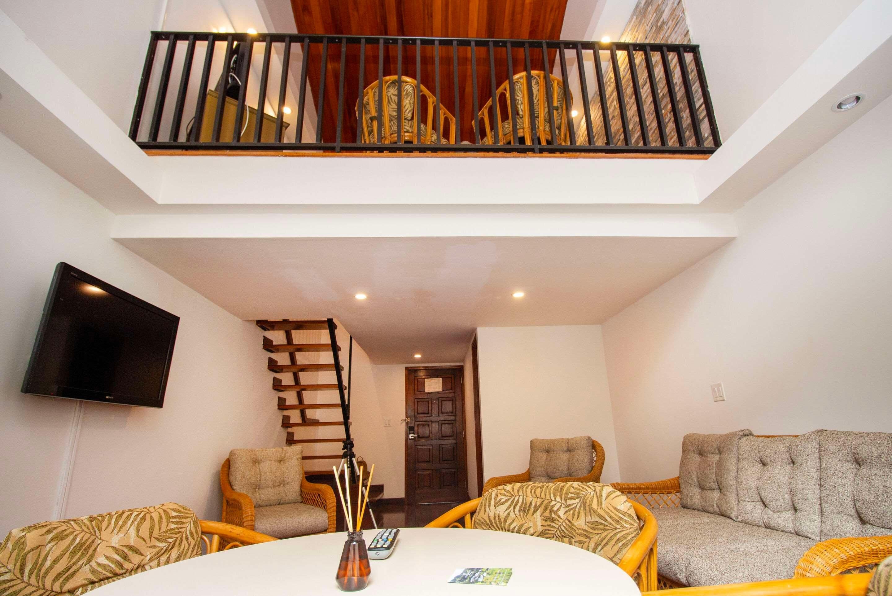 A spacious living area with a high ceiling, a flat-screen TV mounted on the wall, a wooden staircase, and a seating arrangement with two sofas and a coffee table.
