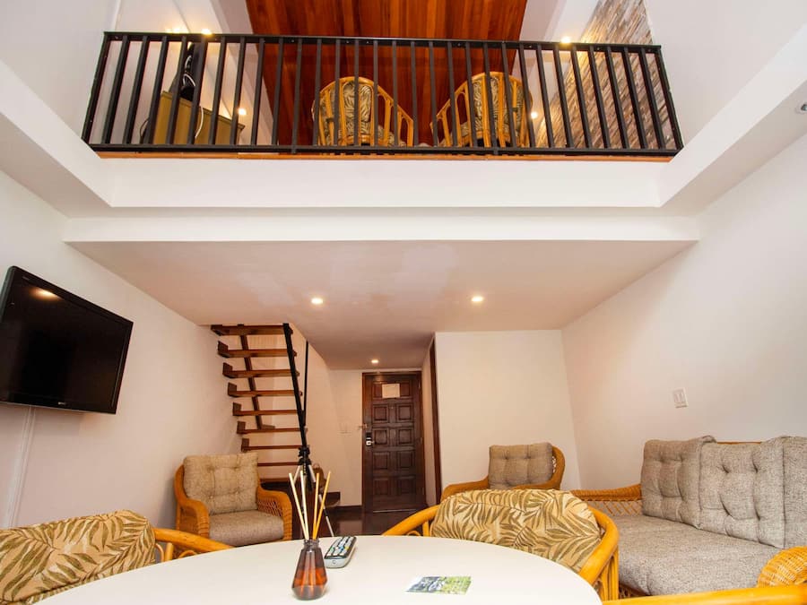 A spacious living area with a high ceiling, a flat-screen TV mounted on the wall, a wooden staircase, and a seating arrangement with two sofas and a coffee table.