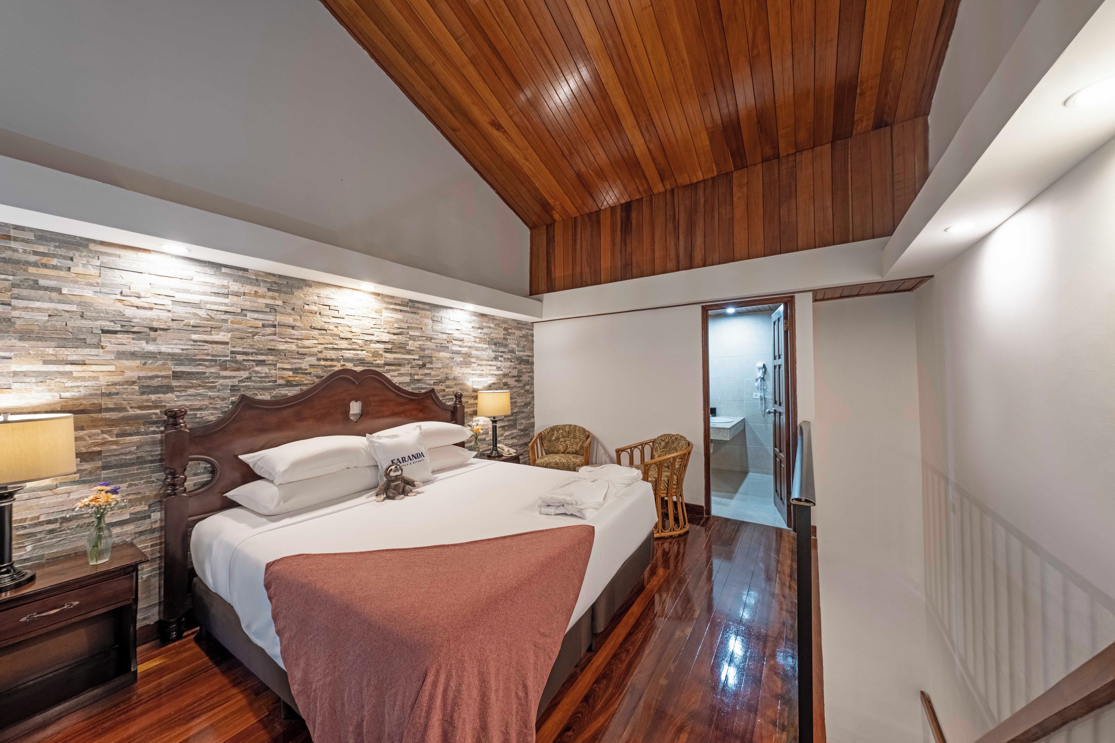 A hotel room with a wooden ceiling, a stone wall, a bed with white linens, a nightstand with a lamp, and a wicker chair.