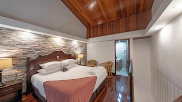 A hotel room with a wooden ceiling, a stone wall, a bed with white linens, a nightstand with a lamp, and a wicker chair.
