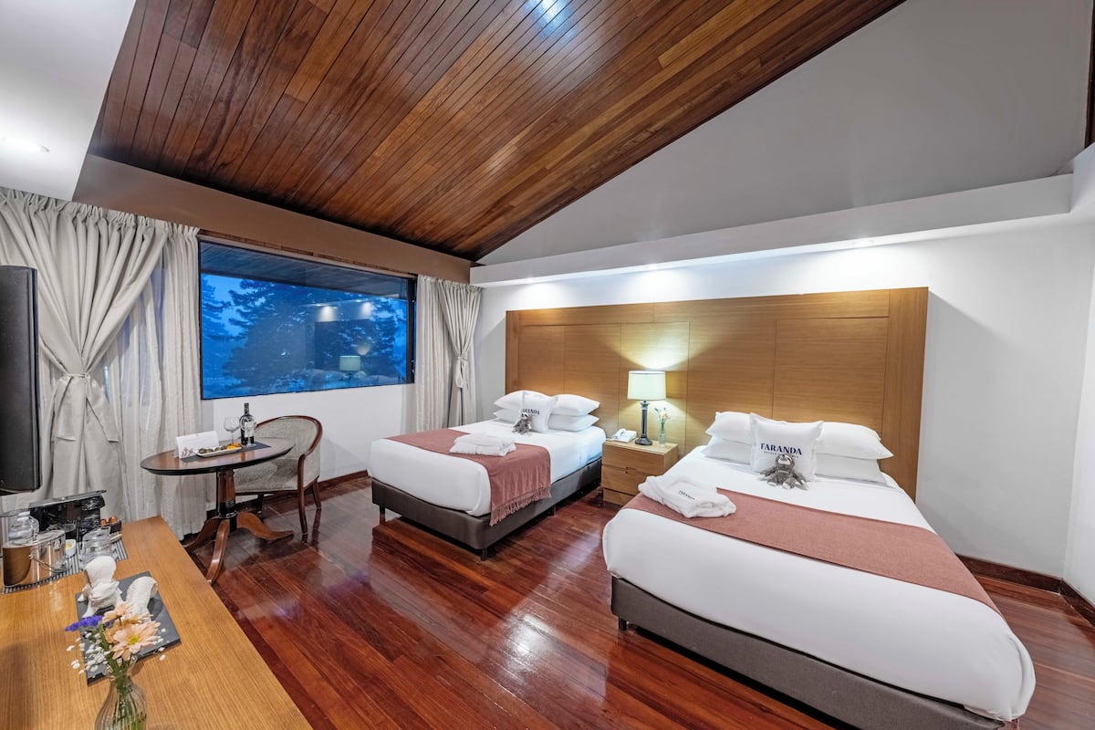 A hotel room with two beds, a wooden ceiling, a large window with curtains, a bedside table, a TV, and a small desk with a vase of flowers.