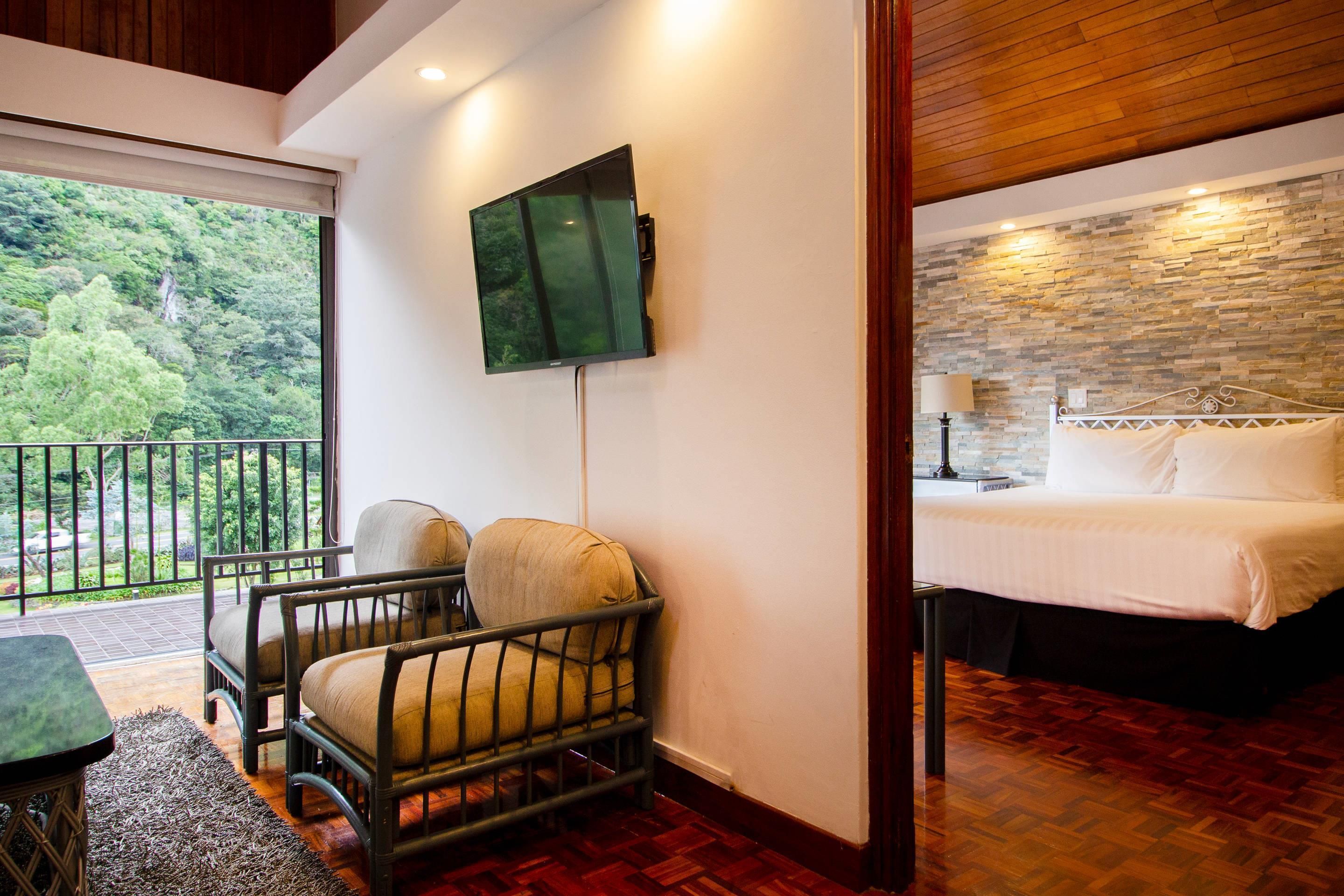 A hotel room with a bed, a chair, a TV mounted on the wall, and a balcony with a view of greenery.