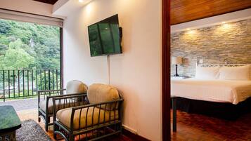 A hotel room with a bed, a chair, a TV mounted on the wall, and a balcony with a view of greenery.