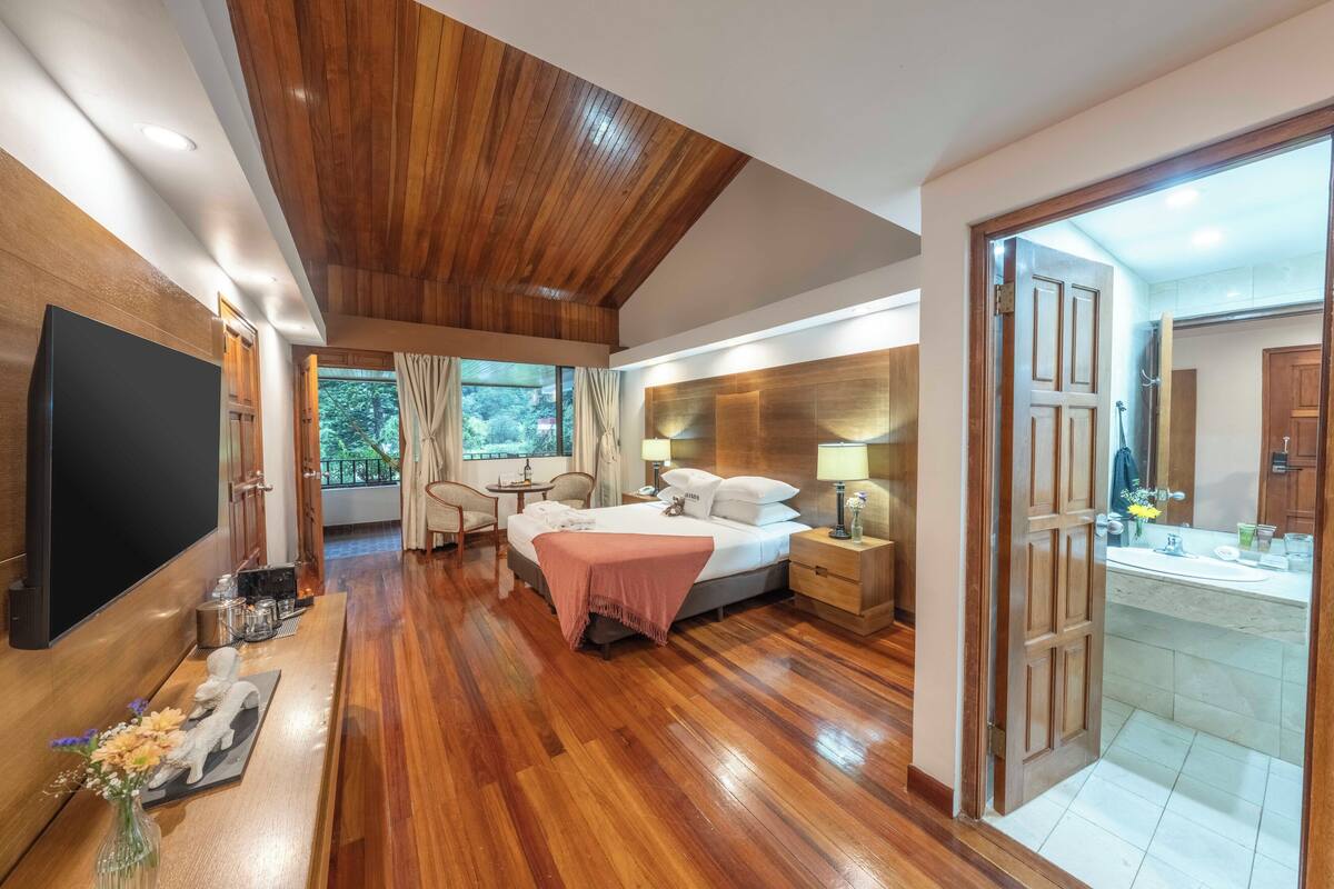A hotel room with a wooden ceiling, a bed with a pink blanket, a TV, a desk with a vase of flowers, and a view of greenery outside.