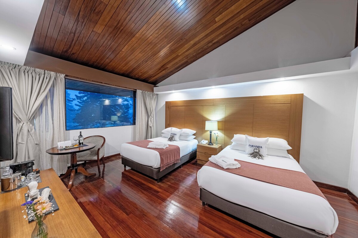 A hotel room with two beds, a wooden ceiling, a large window with curtains, a bedside table, a TV, and a small desk with a vase of flowers.