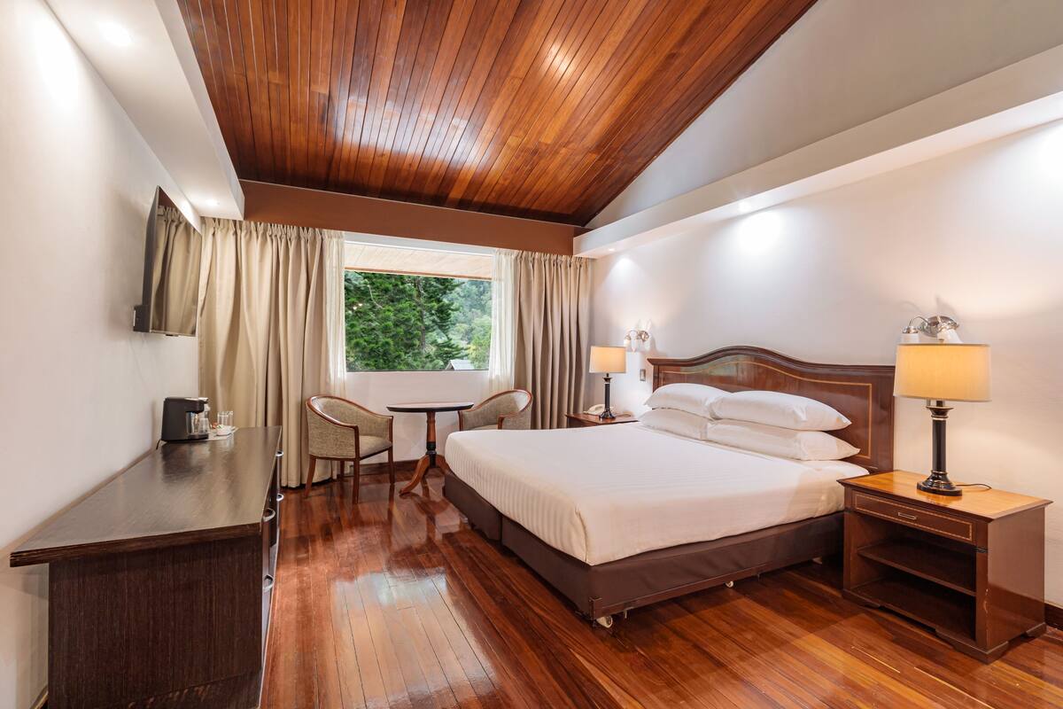 A hotel room with a wooden ceiling, a bed with white linens, a desk with a coffee maker, a chair, a small table, and a nightstand with a lamp.