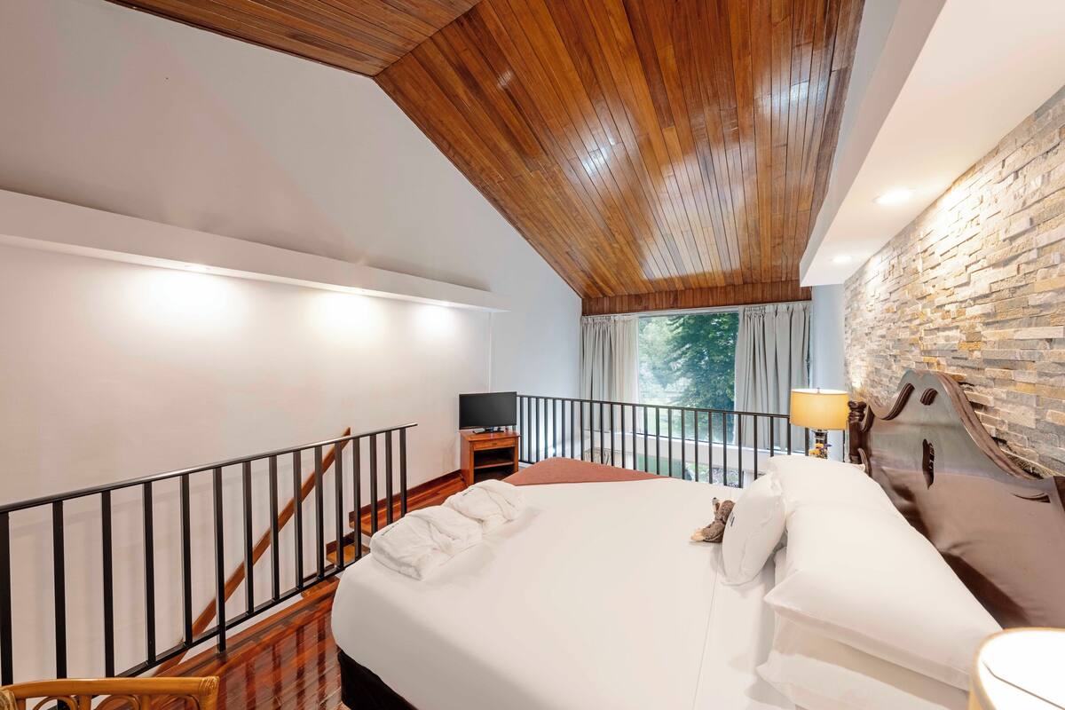 A bedroom with a wooden ceiling, a stone wall, a bed with pillows, a nightstand, a TV, and a window with curtains.
