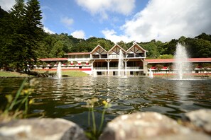A hotel with a water fountain in the foreground, a pool, and a mountainous backdrop.