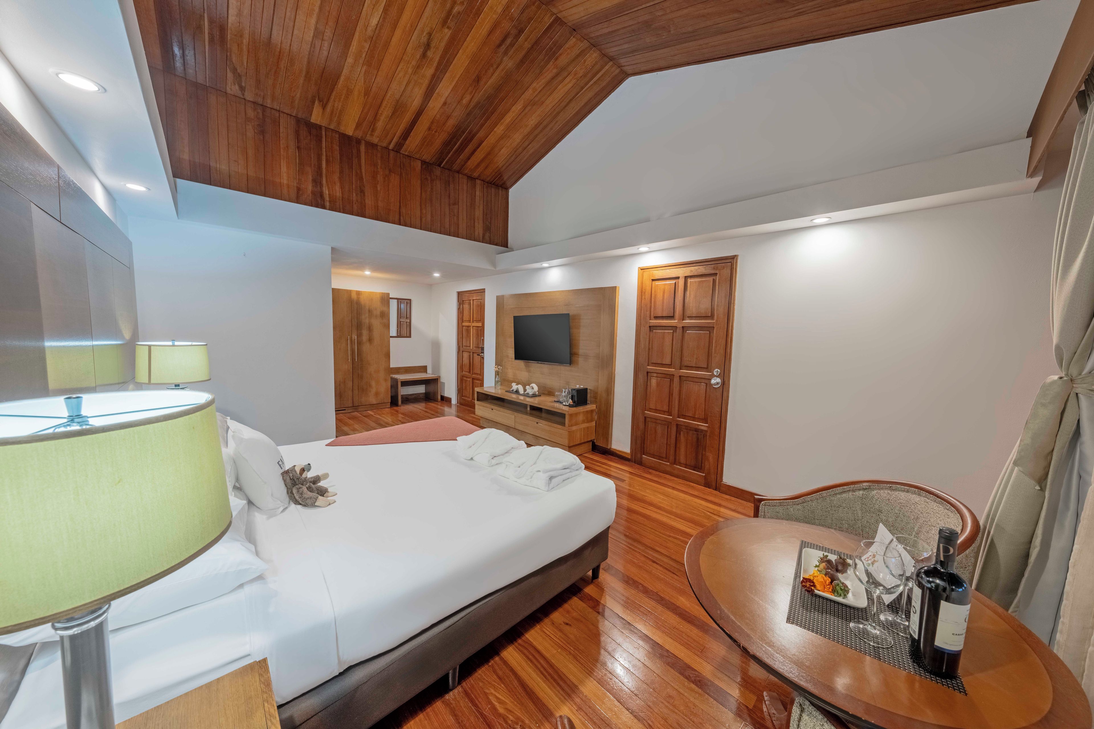 A hotel room with a bed, bedside table, lamp, TV, wooden wardrobe, and a dining area with a table and chairs.
