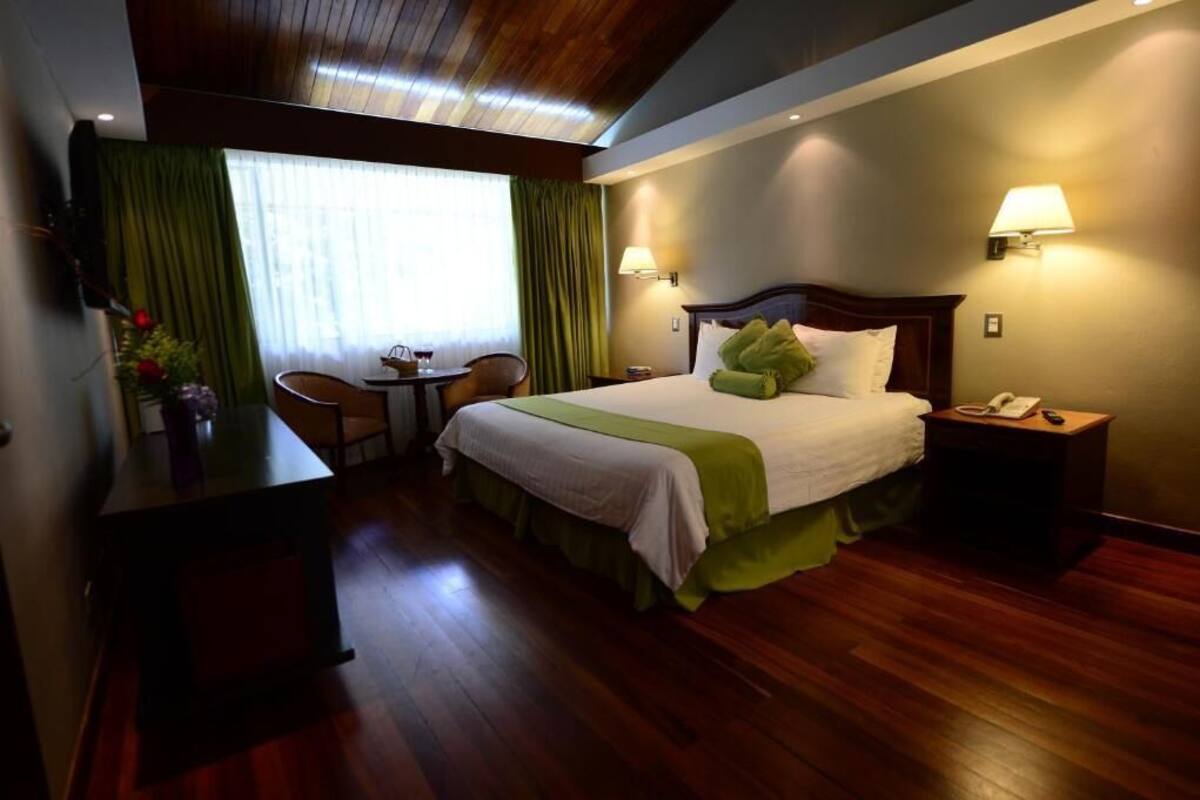 A hotel room with a bed, bedside table, lamp, and a desk with a chair.