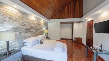 A hotel room with a stone wall, wooden ceiling, a bed with white linens, a bedside table with a lamp, and a TV on the wall.