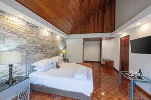 A hotel room with a stone wall, wooden ceiling, a bed with white linens, a bedside table with a lamp, and a TV on the wall.