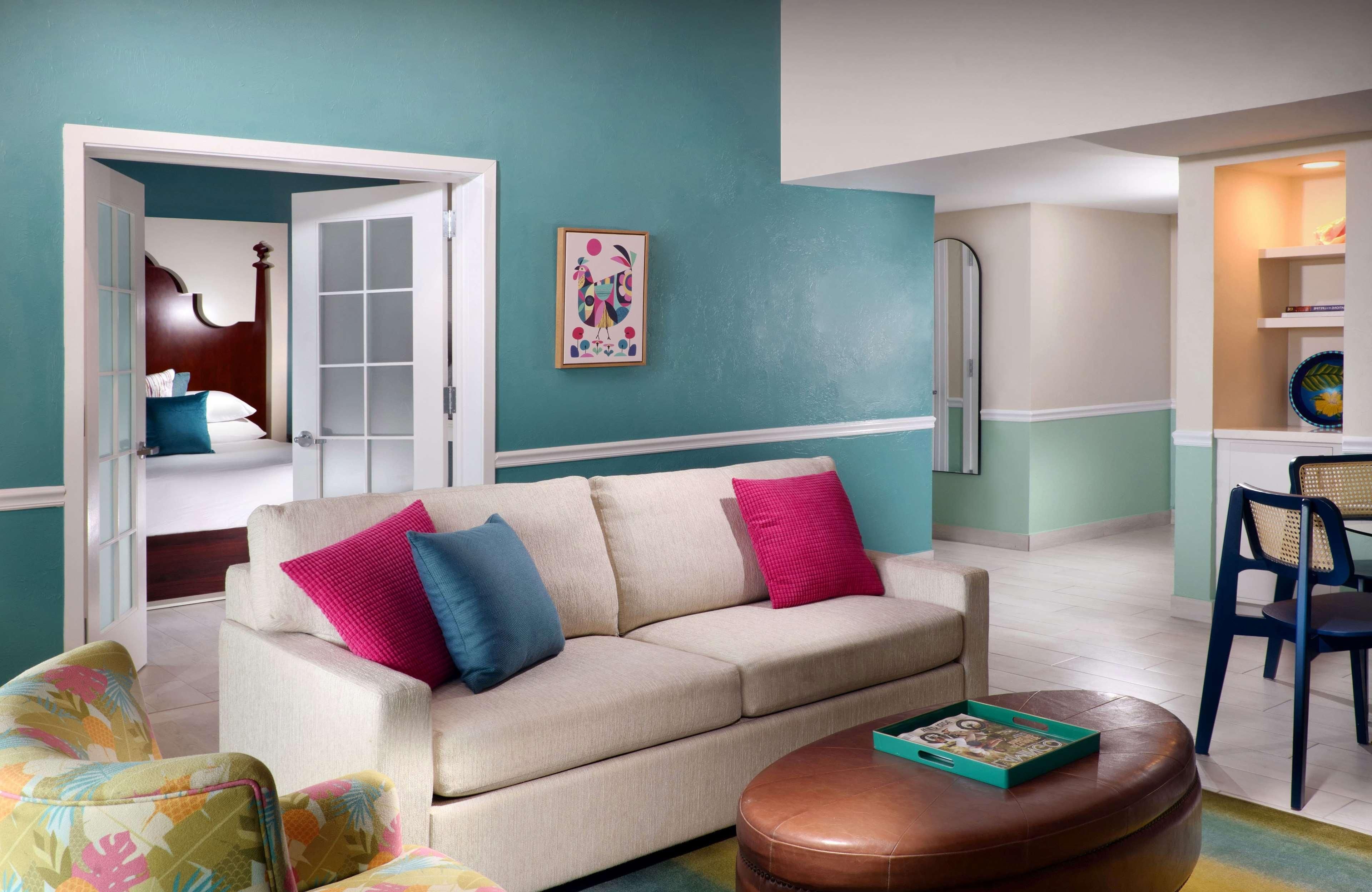 Una sala de estar con sofá color beige, cojines de colores, un reposapiés marrón y una pared verde azulado.
