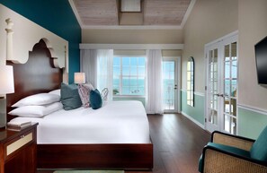 A hotel room with a large bed, a nightstand, a chair, a TV, and a view of the sea through the windows.