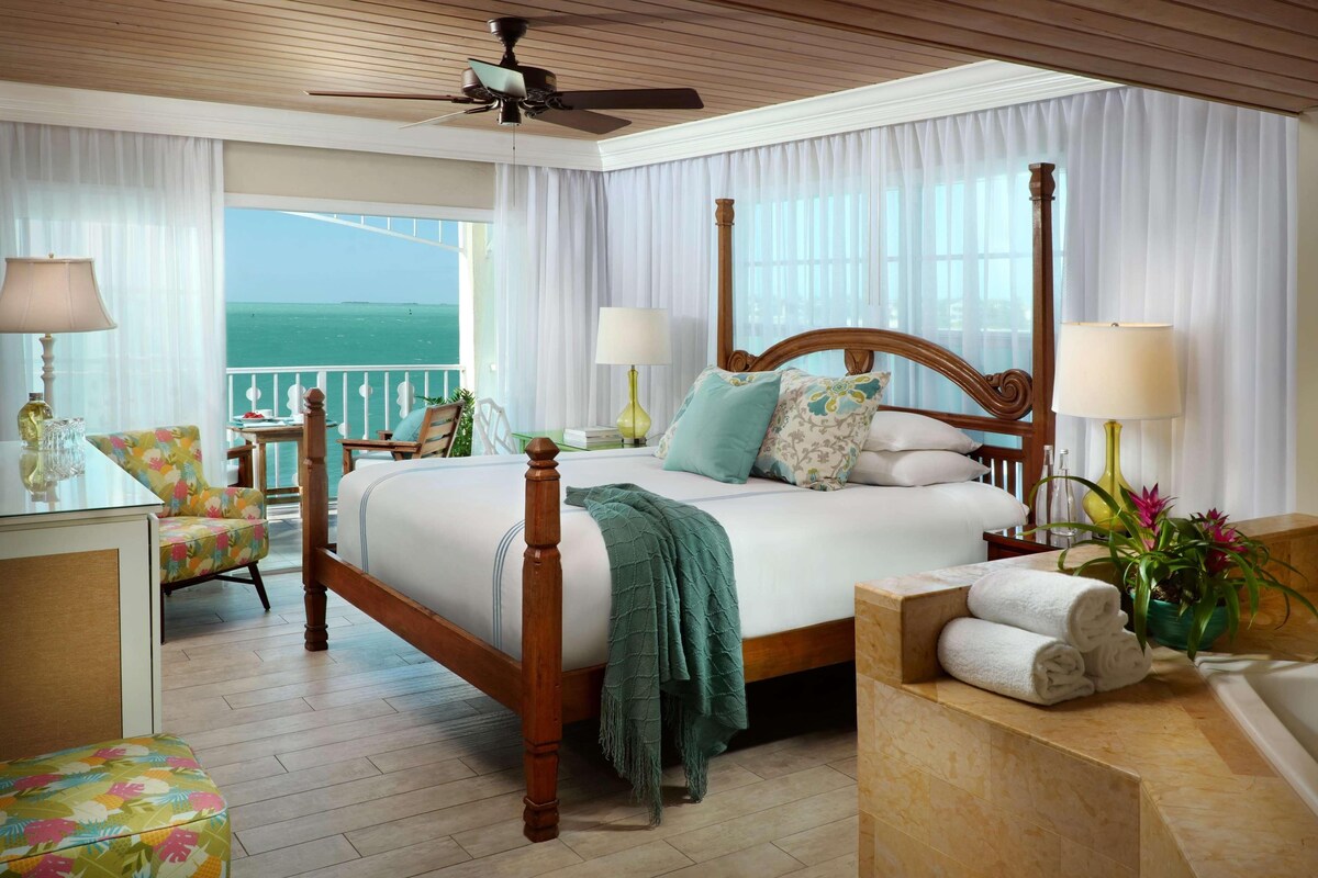 A spacious bedroom with a large bed, a balcony with a view, and a wooden ceiling fan.