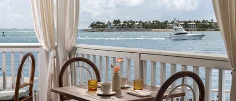 A balcony with a table set for two, overlooking a body of water with a yacht in the distance.