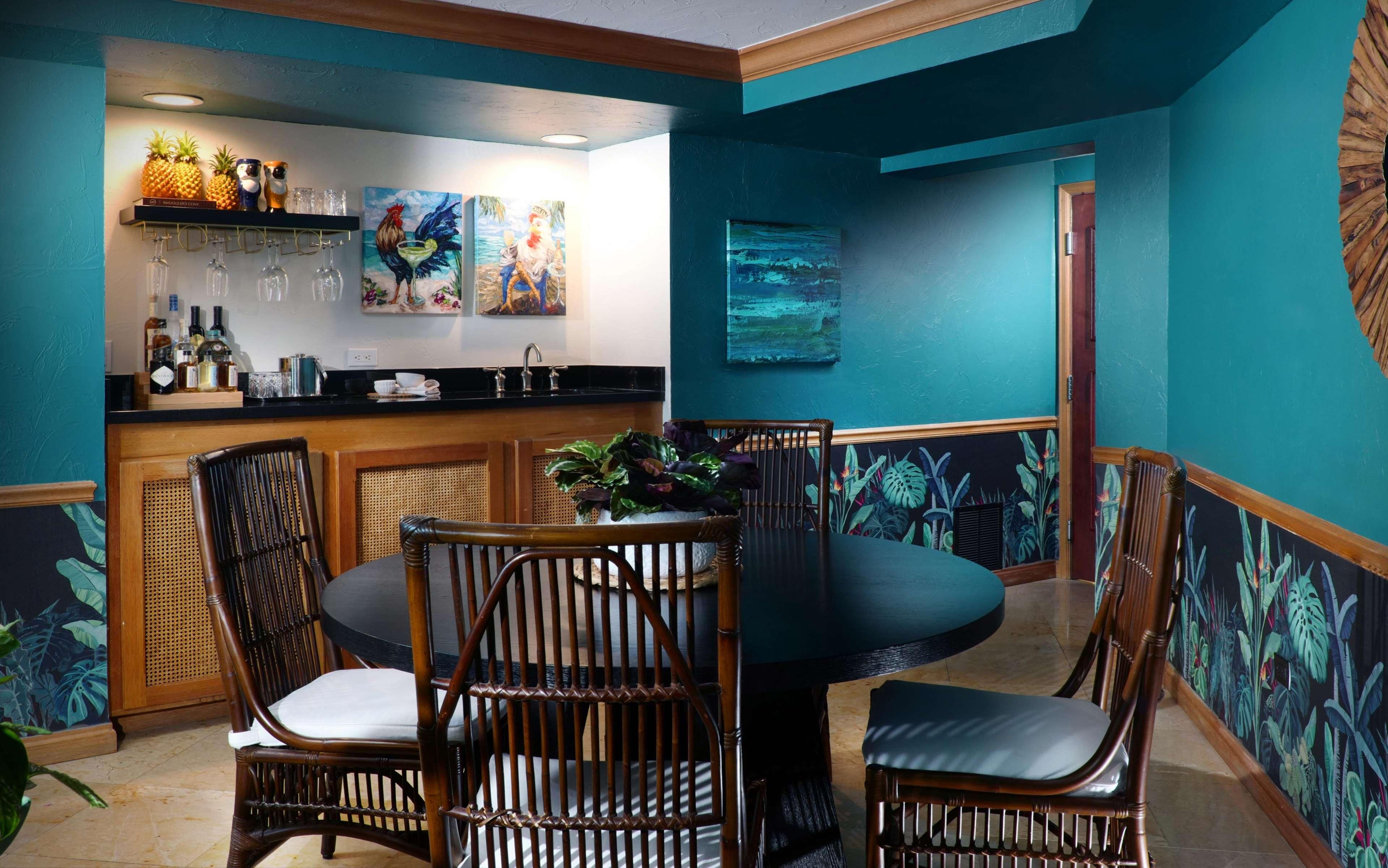 Zona de bar con mostrador, taburetes y una mesa de comedor con sillas. Se observan obras de arte en las paredes y un mural con temática tropical.