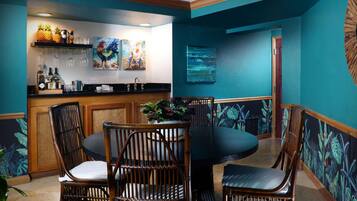 Zona de bar con mostrador, taburetes y una mesa de comedor con sillas. Se observan obras de arte en las paredes y un mural con temática tropical.