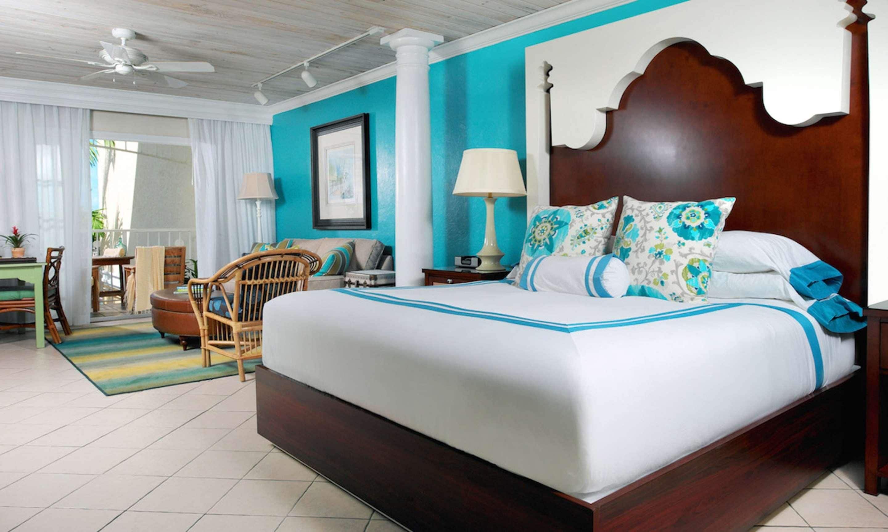 A hotel room with a large bed, a wooden headboard, a white and blue patterned bedspread, a wooden nightstand, a white lamp, and a wicker chair.