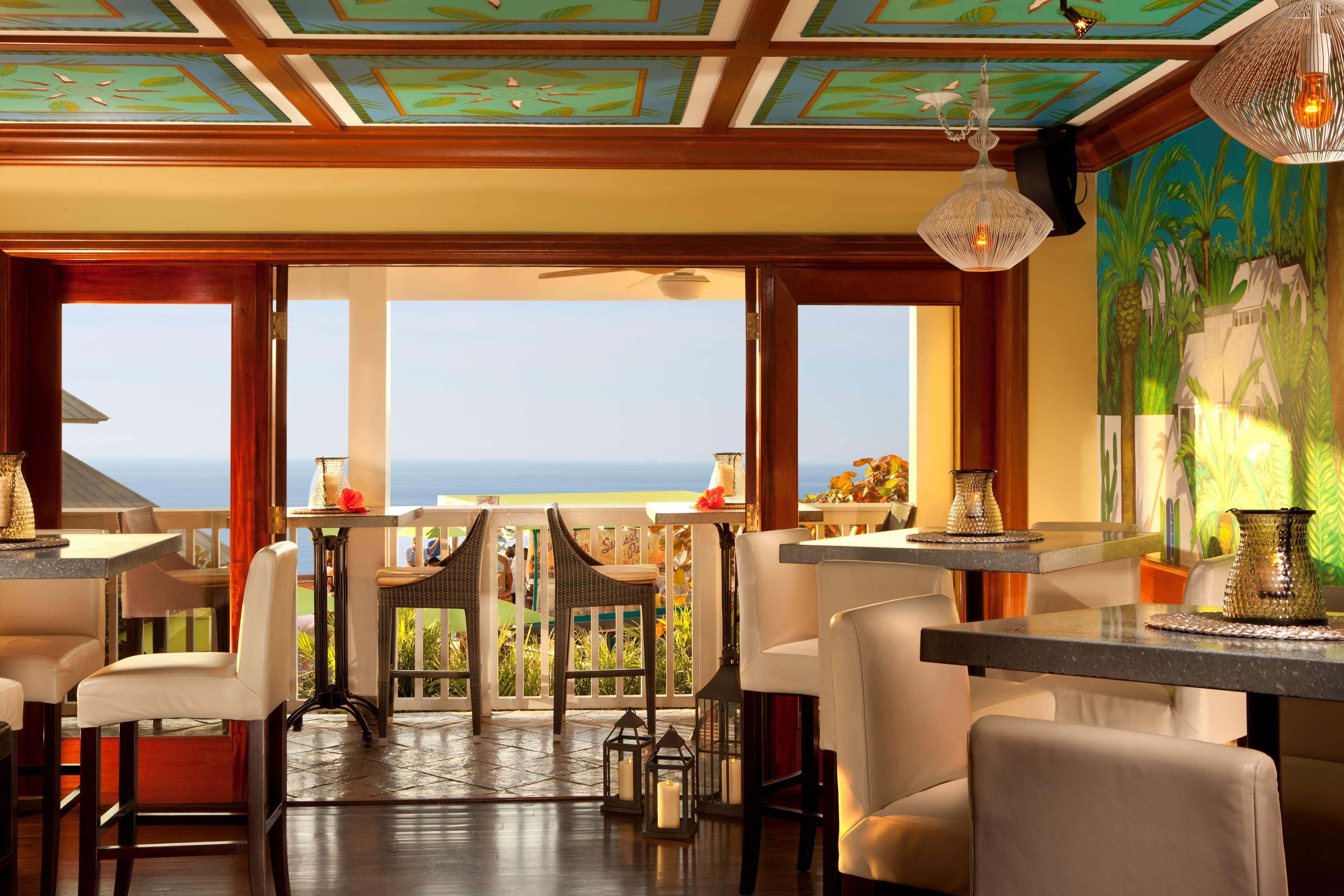 A restaurant with a view of the ocean, featuring a terrace with tables and chairs, a ceiling with a geometric pattern, and a mural on the wall.
