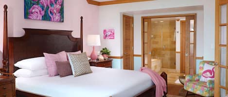 A bedroom with a large bed, wooden headboard, bedside tables, a nightstand, a chair, a painting, and a bathroom with a glass door.