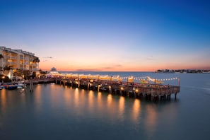A waterfront hotel with a pier extending into calm waters, featuring outdoor seating and a view of the sea.