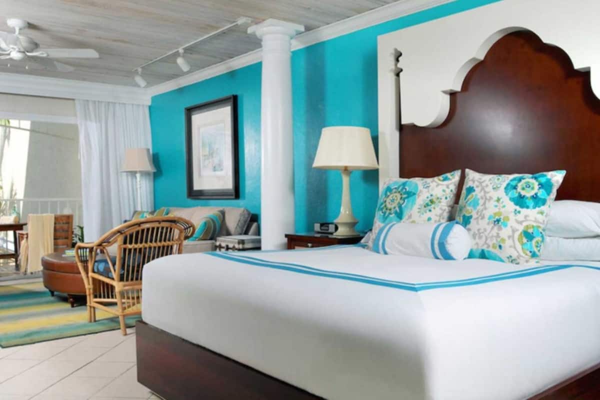 A hotel room with a large bed, a wooden headboard, a nightstand with a lamp, a chair, and a view of the outdoors.